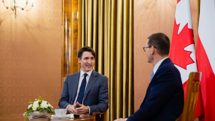 Dwóch mężczyzn siedzących przy stole z kanadyjskimi flagami w tle, w tym premier Justin Trudeau.