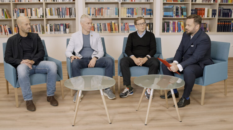 Opis: Czterech mężczyzn siedzi na krzesłach przed biblioteczką.