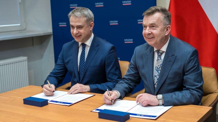 Dwóch mężczyzn w strojach biznesowych siedzi przy stole i uśmiecha się podczas podpisywania dokumentów z flagami w tle.