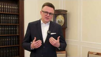 Mężczyzna w ciemnym garniturze i okularach gestykuluje rękami podczas rozmowy w eleganckim pokoju.