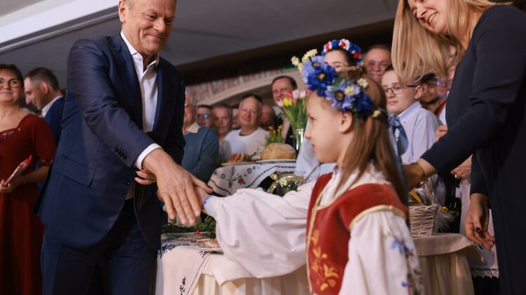 Mężczyzna w garniturze podaje rękę młodej dziewczynie w tradycyjnym stroju podczas wydarzenia publicznego, na oczach widzów.