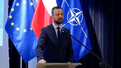 Mężczyzna wygłaszający przemówienie na podium z flagami UE i NATO w tle.