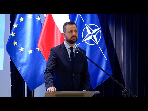 Mężczyzna wygłaszający przemówienie na podium z flagami UE i NATO w tle.