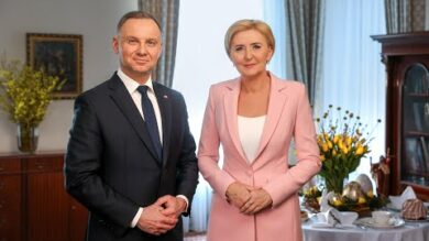 Dwie osoby, mężczyzna i kobieta, stoją formalnie w pokoju ze stołem nakrytym do posiłku w tle.