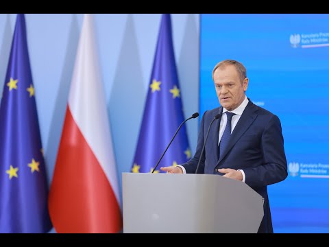 Mężczyzna wygłaszający przemówienie na podium, na tle Unii Europejskiej i polskich flag.