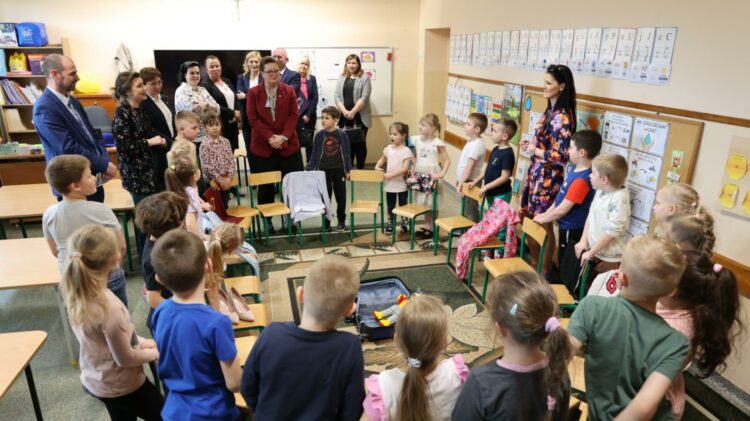 Dzieci stojące w klasie twarzą do dorosłych, prawdopodobnie podczas szkolnego wydarzenia lub prezentacji.