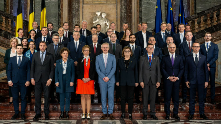 Zdjęcie grupowe formalnie ubranych osób na oficjalnym wydarzeniu, z flagami w tle wskazującymi kontekst europejski.