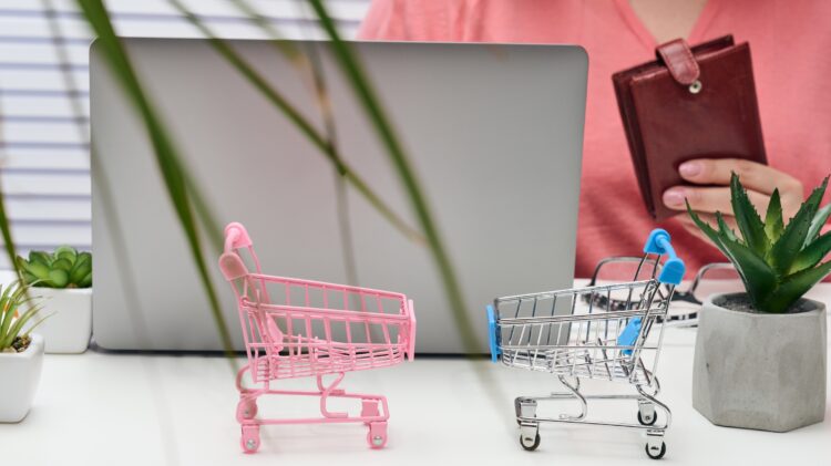 Osoba siedząca za laptopem z miniaturowymi wózkami na biurku, symbolizującymi zakupy online.
