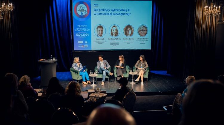 Czterech panelistów siedzi na scenie podczas konferencji, omawiając sztuczną inteligencję w komunikacji, przed publicznością w słabo oświetlonym pokoju.