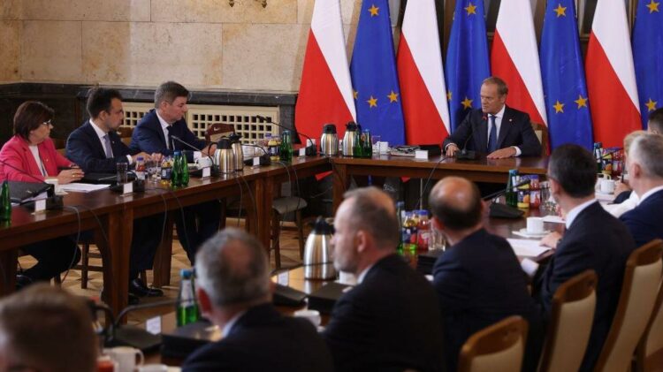 Grupa urzędników w strojach formalnych siedzi wokół dużego stołu w sali konferencyjnej, na tle flag narodowych i Unii Europejskiej.