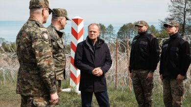 Czterech mężczyzn, w tym dwóch w mundurach wojskowych, prowadzi poważną dyskusję przy znaku granicznym, na tle zalesionego terenu i spokojnego morza.