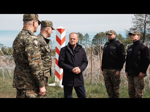 Czterech mężczyzn, w tym dwóch w mundurach wojskowych, prowadzi poważną dyskusję przy znaku granicznym, na tle zalesionego terenu i spokojnego morza.