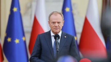 Za mikrofonem stoi mężczyzna w garniturze, w tle flagi Unii Europejskiej i Polski.