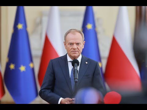 Za mikrofonem stoi mężczyzna w garniturze, w tle flagi Unii Europejskiej i Polski.