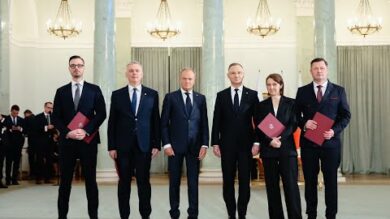 Pięciu urzędników stojących w formalnym rzędzie w eleganckiej sali, trzymających czerwone teczki podczas ceremonii.