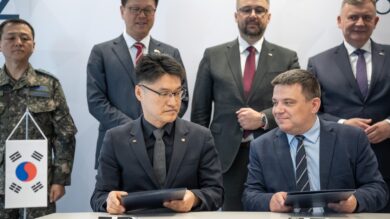 Dwóch urzędników z Korei Południowej i Słowacji siedzi przy stole i podpisuje dokumenty, co obserwuje w tle grupa personelu wojskowego i biznesmenów.