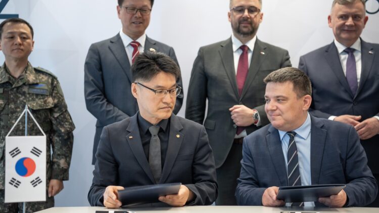 Dwóch urzędników z Korei Południowej i Słowacji siedzi przy stole i podpisuje dokumenty, co obserwuje w tle grupa personelu wojskowego i biznesmenów.