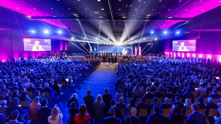 Duża publiczność na konferencji, skupiona na scenie z niebieskim i fioletowym oświetleniem, na której gromadzi się kilka osób.