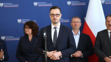 Mężczyzna w garniturze stojący na podium z mikrofonem, w otoczeniu innych osób, na tle z oficjalnymi logo.