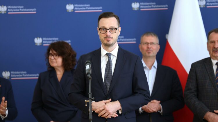 Mężczyzna w garniturze stojący na podium z mikrofonem, w otoczeniu innych osób, na tle z oficjalnymi logo.