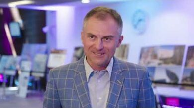 Mężczyzna w niebieskim garniturze w kratkę stoi w pomieszczeniu z uśmiechem. W tle znajduje się kilka plakatów i rozmyta scena wskazująca miejsce wydarzenia lub prezentacji.