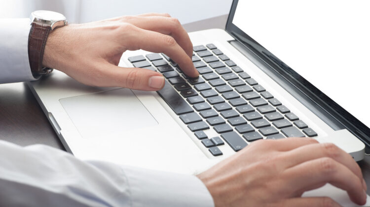 Osoba nosząca zegarek i koszulę z długim rękawem pisze na klawiaturze laptopa stojącego na biurku.