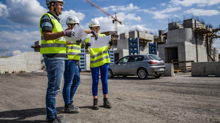 Trzej pracownicy budowlani w sprzętach ochronnych i kaskach przeglądają plany w miejscu pracy, na tle częściowo wybudowanych budynków i zaparkowanego samochodu.
