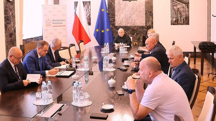 Grupa osób w formalnej sali konferencyjnej siedzi przy długim stole z ułożonymi dokumentami, butelkami wody i szklankami. W tle flagi Polski i Unii Europejskiej.