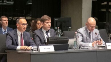 Przy stole konferencyjnym z mikrofonami siedzi kilka osób w strojach formalnych. Na tabliczkach znamionowych przed niektórymi delegatami widniały napisy „POLSKA” i „MAGYARORSZÁG.