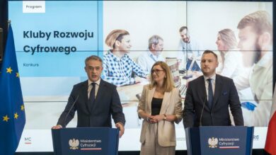 Podczas konferencji prasowej na podium stoją trzy osoby, każda ubrana w profesjonalny strój. Za nimi znajduje się duży ekran, na którym wyświetlany jest napis „Klub Rozwoju Cyfrowego”. W tle dwie flagi narodowe.