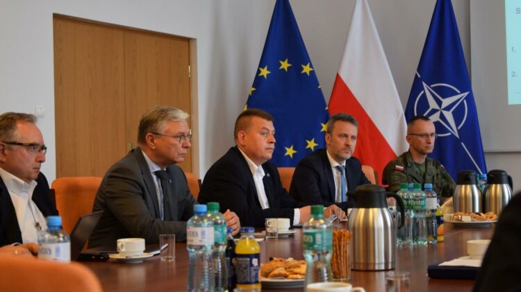 Grupa mężczyzn w garniturach i żołnierz siedzą przy stole konferencyjnym, a za nimi flagi UE, Polski i NATO. Stół zastawiony jest napojami, szklankami, filiżankami i przekąskami.