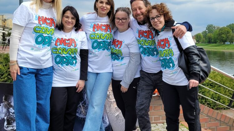 Grupa sześciu osób stojących na zewnątrz uśmiecha się do kamery. Mają na sobie białe T-shirty z kolorowymi tekstami i grafikami. W tle widać rzekę i drzewa.