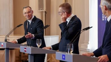 Trzej mężczyźni stoją na podium w formalnym otoczeniu, każdy z mikrofonem i szklanką wody. Jeden mężczyzna gestykuluje podczas mówienia, a drugi poprawia słuchawkę. Flagi i mikrofony są widoczne.