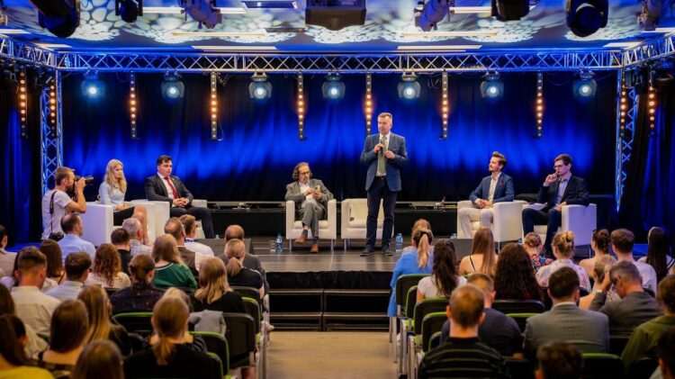 Mężczyzna stoi i przemawia podczas dyskusji panelowej przed publicznością. Sześciu siedzących panelistów znajduje się na scenie w tle niebieskiego oświetlenia i świateł scenicznych.