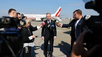 Mężczyzna w garniturze przemawia do wielu mikrofonów trzymanych przez reporterów na płycie lotniska, z samolotem w tle.