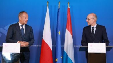 Dwóch mężczyzn w garniturach stoi na podium z mikrofonami przed flagami, w tym polską, unijną i luksemburską, na niebieskim tle.