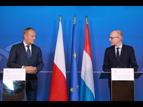 Dwóch mężczyzn w garniturach stoi na podium z mikrofonami przed flagami, w tym polską, unijną i luksemburską, na niebieskim tle.