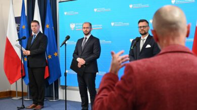 Trzej mężczyźni w garniturach stoją podczas konferencji prasowej przed mikrofonami i flagami, w tym polską i unijną. Osoba z łysą głową wskazuje na nie, odwrócona tyłem do kamery.