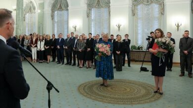 Pokój wypełniony ludźmi, pośrodku dwie kobiety trzymające bukiety kwiatów i mężczyzna przemawiający na podium. W pomieszczeniu znajdują się łukowe okna z zasłonami i żyrandolami.