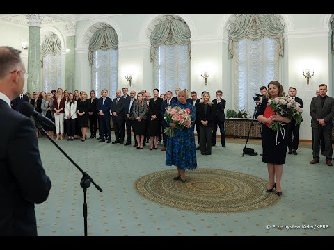 Pokój wypełniony ludźmi, pośrodku dwie kobiety trzymające bukiety kwiatów i mężczyzna przemawiający na podium. W pomieszczeniu znajdują się łukowe okna z zasłonami i żyrandolami.