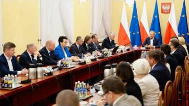 Grupa osób w strojach formalnych siedzi wokół dużego stołu konferencyjnego i pogrąża się w dyskusji. W tle widoczne flagi narodowe. Woda butelkowana i mikrofony są na stole.