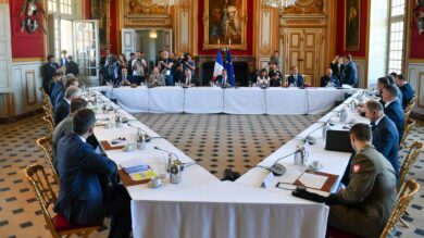 Duże spotkanie z udziałem około 20 osób siedzących przy stole w kształcie litery U w bogato zdobionej sali z żyrandolami i obrazami. Mówca stoi na podium pod flagą, a fotografowie i inne osoby obserwują.