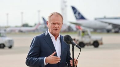 Mężczyzna w garniturze przemawia do mikrofonu podczas imprezy plenerowej na płycie lotniska, na tle kilku samolotów widocznych.