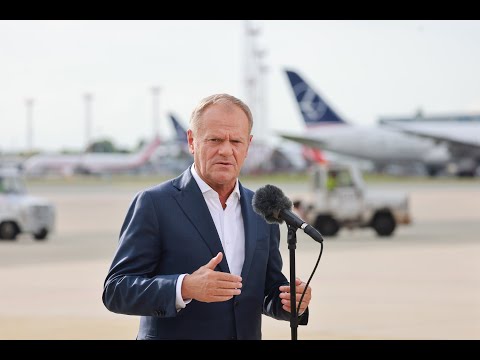 Mężczyzna w garniturze przemawia do mikrofonu podczas imprezy plenerowej na płycie lotniska, na tle kilku samolotów widocznych.