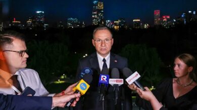 Mężczyzna w garniturze przemawia nocą przy stojaku mikrofonowym na świeżym powietrzu, otoczony przez kilku reporterów trzymających mikrofony, na tle oświetlonej panoramy miasta.