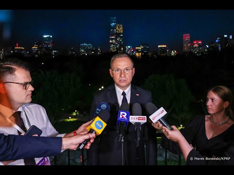 Mężczyzna w garniturze przemawia nocą przy stojaku mikrofonowym na świeżym powietrzu, otoczony przez kilku reporterów trzymających mikrofony, na tle oświetlonej panoramy miasta.
