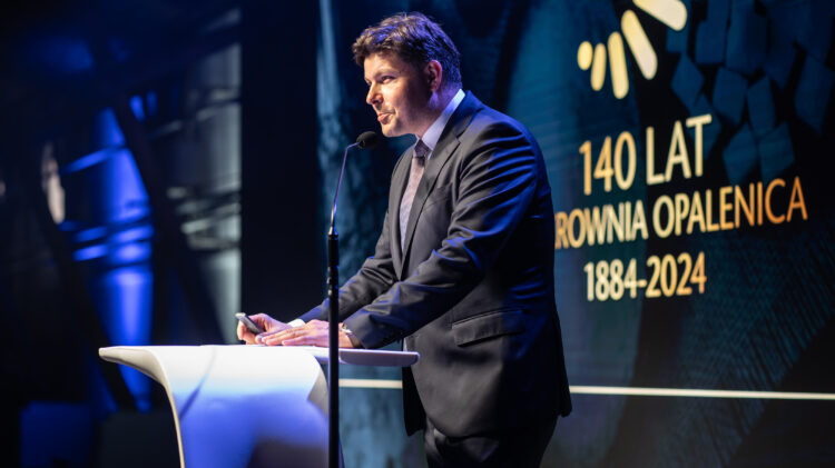 Mężczyzna w garniturze przemawia na podium podczas wydarzenia. W tle widnieje tekst: „140 LAT CUCROWNIA OPALENICA 1884-2024.