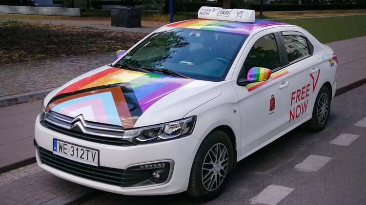 Biała taksówka Citroën z logo Free Now i tęczową flagą z wzorem w jodełkę na masce i dachu, zaparkowana na ulicy miejskiej.