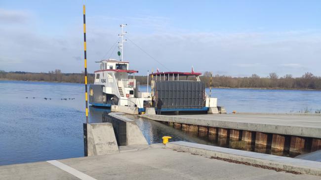 Prom zacumowany przy betonowym molo nad spokojną rzeką z czystym niebem w tle. Stacja dokująca posiada żółto-czarną barierkę zabezpieczającą. Drzewa rosną na przeciwległym brzegu rzeki.