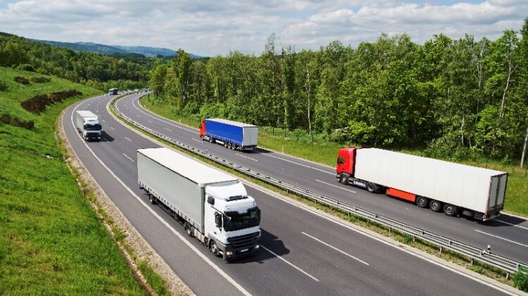Kilka półciężarówek jedzie podzieloną autostradą otoczoną zielenią i drzewami, pod niebem z rozproszonymi chmurami.